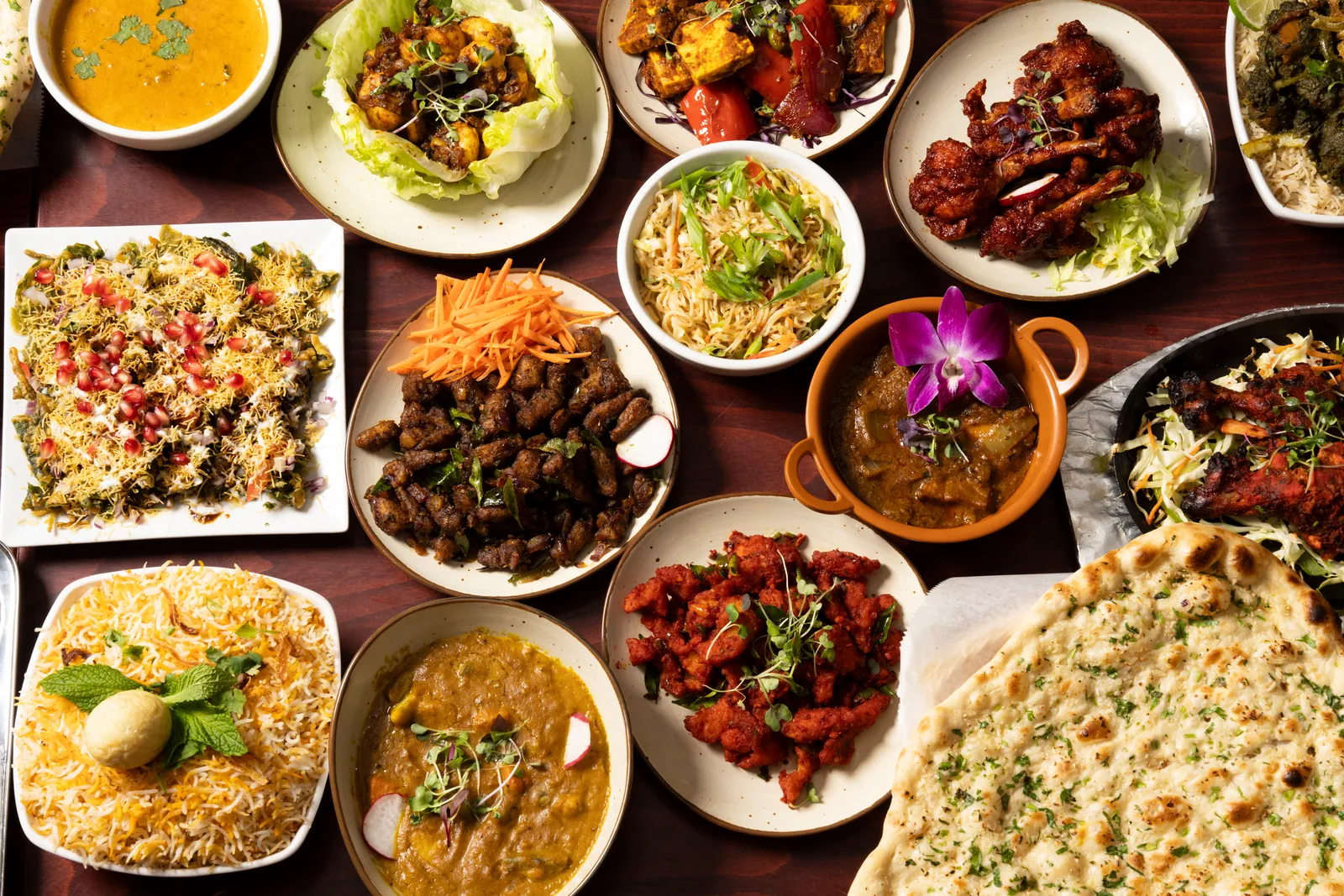 Overhead spread of Indian dishes on a dark wood table: biryani, dal, curries, chili chicken, chaat and garlic naan
