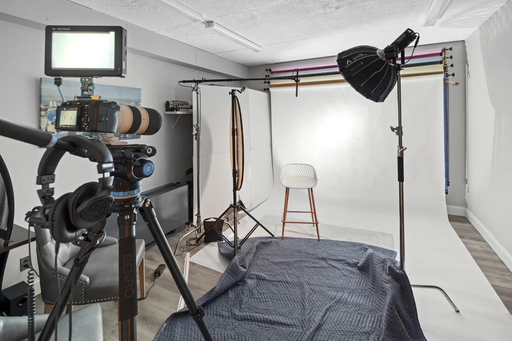 Studio shooting bay with a white seamless backdrop, stool, softbox on a boom arm, reflector, and a camera on a tripod feeding a monitor