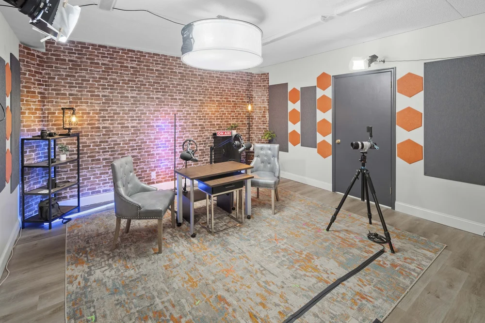 Podcast and interview room with an exposed brick wall, two upholstered chairs either side of a table with boom microphones, an ON AIR sign, and acoustic panels