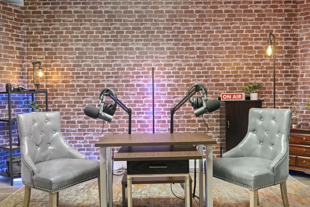 Two-seat podcast set photographed head-on: boom microphones and headphones over a wooden table against a lit brick wall
